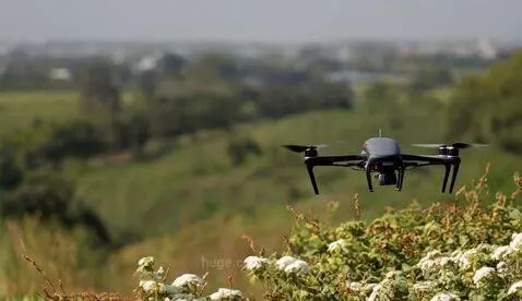 Gray drone with four propellers flying over a green landscape with bushes and rolling hills.