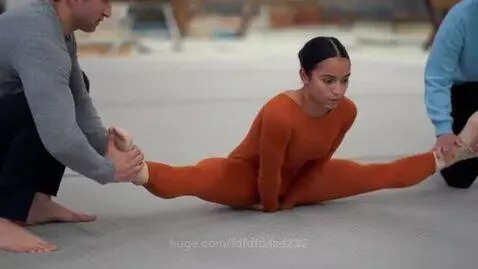 Gymnast on back on floor, assisted into a full split by two people holding her feet.