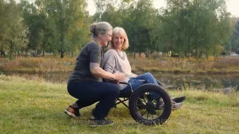 Two elderly women smiling, one in a wheelchair, the other kneeling beside her, holding hands outdoors by water.