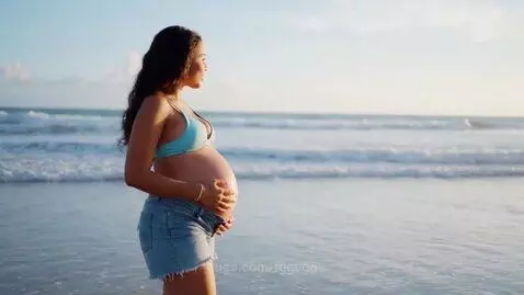 Pregnant woman with dark hair wearing a bikini top and denim shorts, standing on a beach and holding her belly, ocean in background.