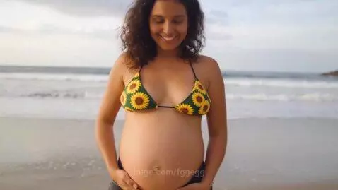 Pregnant woman with curly dark hair wearing a sunflower bikini top and black denim shorts on a sandy beach, holding her belly.