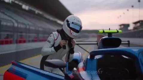 Race car driver in white suit and helmet entering a blue and red race car on a track.