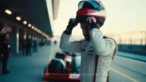 Race car driver in a red and white suit putting on his helmet on a race track, with car number 277 visible.