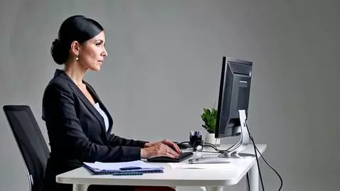 Professional woman with dark hair in a bun, wearing a business suit, typing on a keyboard at a modern white desk in an office.