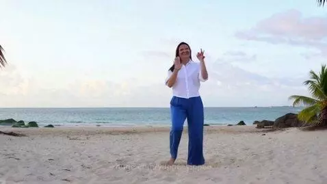 Woman with dark hair in white shirt and blue pants dancing joyfully on a sandy beach with the ocean in the background.