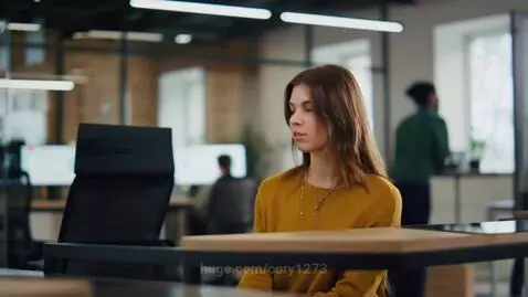 Woman with long brown hair at a desk in a modern office, with blurred colleagues and computers in the background.