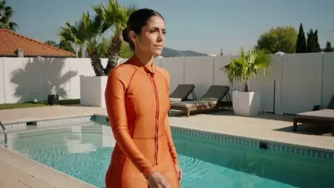 Woman in a bright orange hydrasuit walking beside a swimming pool on a sunny day with palm trees in the background.
