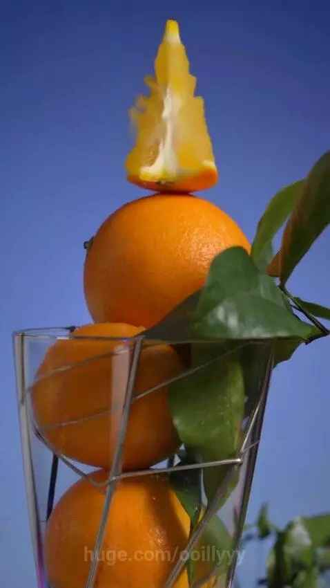 A clear glass vase filled with a stack of fresh oranges and green leaves, with an orange slice on top, spinning against a blue sky.