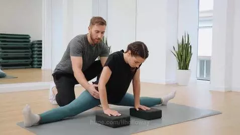 Man assisting woman in middle split stretch with yoga blocks in a bright studio.