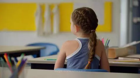 Girl with light brown braid wearing a blue and white striped racerback top, sitting at a desk in a classroom.
