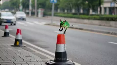 A small green creature with wings jumps off a traffic cone as a car passes by.