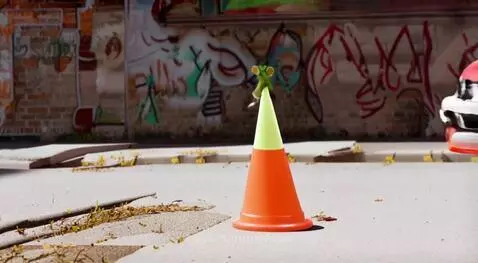 Animated green mouse sits on a traffic cone as a car speeds past.
