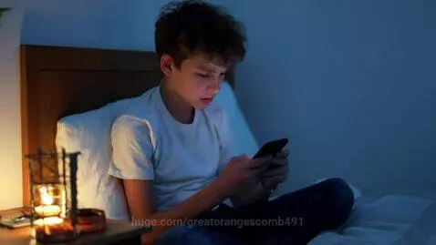 Teenage boy with curly brown hair looking sadly at his smartphone in a dimly lit room.