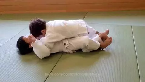 Two young boys in white judo uniforms grappling on a green mat, one boy on his back, the other on top with legs intertwined.