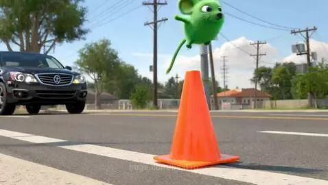 A small green mouse with large ears jumps onto an orange traffic cone as a car speeds past on a sunny day.