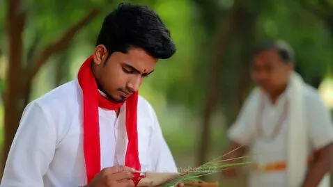 Young Indian man in white kurta-pajama and red scarf writing on a palm-leaf manuscript.