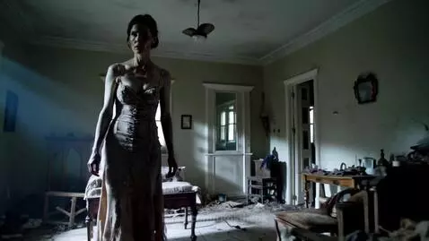 Zombie woman with glowing blue eyes in a torn wedding dress, standing in a dark, dilapidated room with debris.