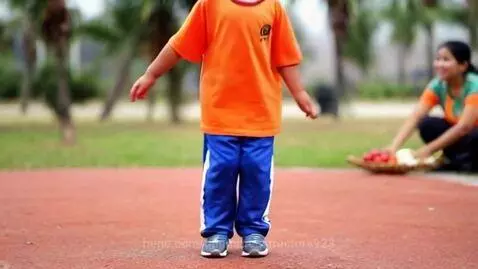 Young child in orange t-shirt and blue pants jumping on a red surface outdoors with green grass and trees in the background.