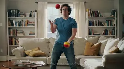 Man in blue shirt and jeans attempting to juggle, then falling backward onto a couch.