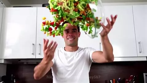 Man in kitchen smiling with salad ingredients flying around him, lettuce on his forehead.