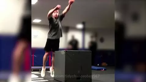 Man in grey t-shirt and black shorts falling backward after missing a box jump in a gym.