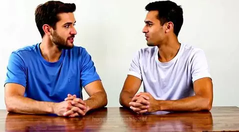 Two young men, one with a beard, sitting at a wooden table, smiling at the camera against a white background.