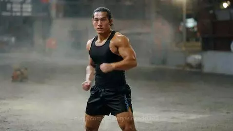 Muscular male fighter with long dark hair in a black tank top and shorts, performing martial arts movements in a dusty indoor gym.