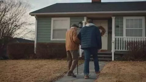 An older man with a cane walks with a younger man up a stone path towards a green house.