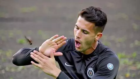 Arsenal player in black shirt swatting at a wasp near his face outdoors.