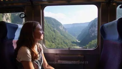 Woman with long brown hair looking out a train window at green mountains and a valley.