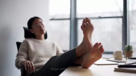 Asian woman in office chair with feet resting on desk, wearing a sweater and pants.