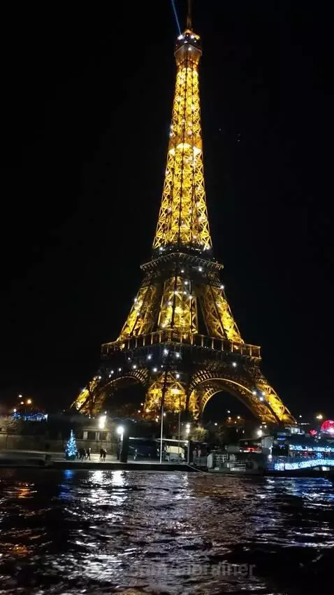 The Eiffel Tower illuminated at night with golden and white lights, reflecting on the Seine River with people on the banks.