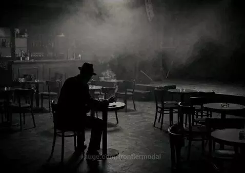 Black and white image of a man in a hat smoking in a dimly lit, smoky bar with a drink and jazz music.