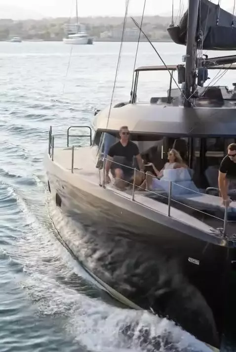 Couple enjoying a sunny day on a black and gold catamaran, with one person doing squats and the other relaxing.