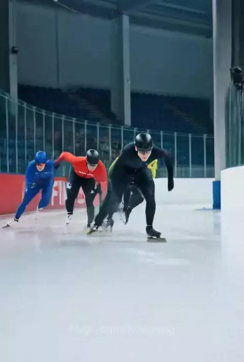 Speed skaters in aerodynamic suits and helmets racing on an indoor ice rink.