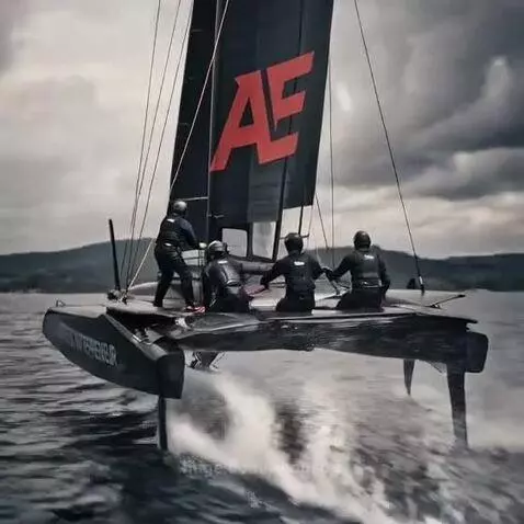 Black catamaran with 'ATHLETIC ENTREPRENEUR' and red 'AE' logo hydrofoiling rapidly across water under cloudy sky.