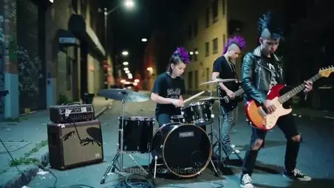 Three teenagers with mohawks performing punk rock music on a street at night with graffiti in the background.