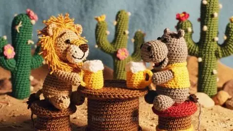 Crocheted lion and hippo figures sitting at a table in a desert with cacti, holding small mugs and smiling.