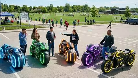 Five adults stand next to futuristic motorcycles with glowing lights in a parking lot.