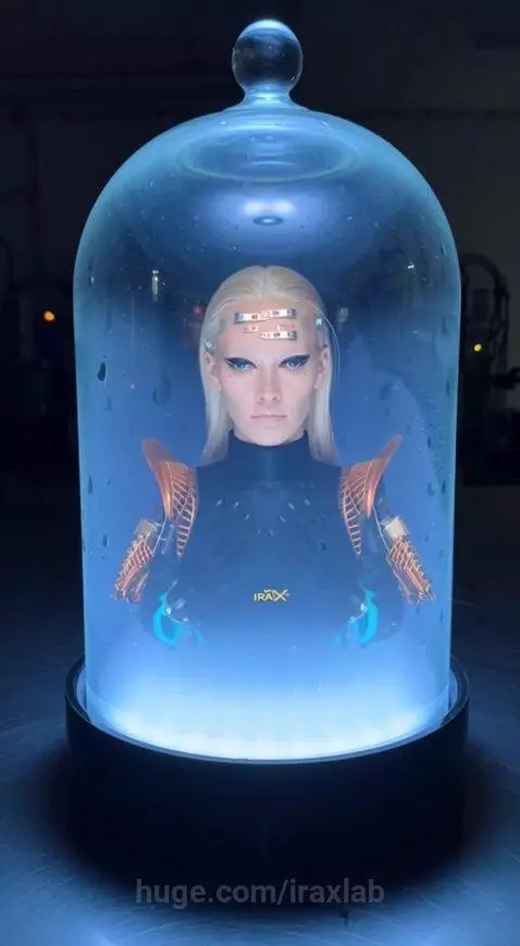 Futuristic blonde woman with metallic accents in a glass dome filled with blue glowing mist, wearing a black garment with 'IRAX'.