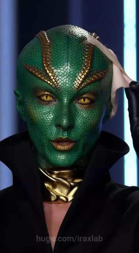 Close-up of a woman with green, scaly skin, gold forehead markings, and yellow slitted eyes, identifying as reptilian.
