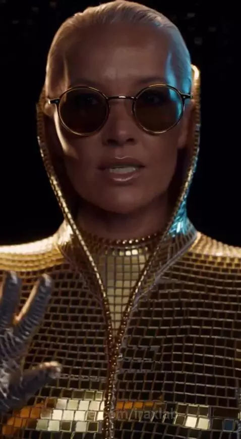 Woman with light hair wearing a gold mosaic hooded jacket and gloves, with round sunglasses, against a dark background.