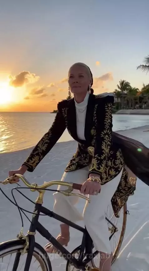 Woman in elegant outfit riding a bicycle with gold accents on a beach at sunset.