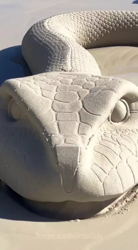 Woman in a black and gold patterned robe on a white sand beach, interacting with a large sand snake sculpture.