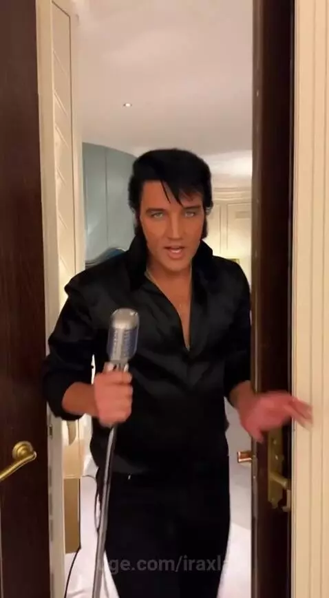 Elvis impersonator in a black satin shirt singing into a vintage microphone, with a woman in a white suit beside him.