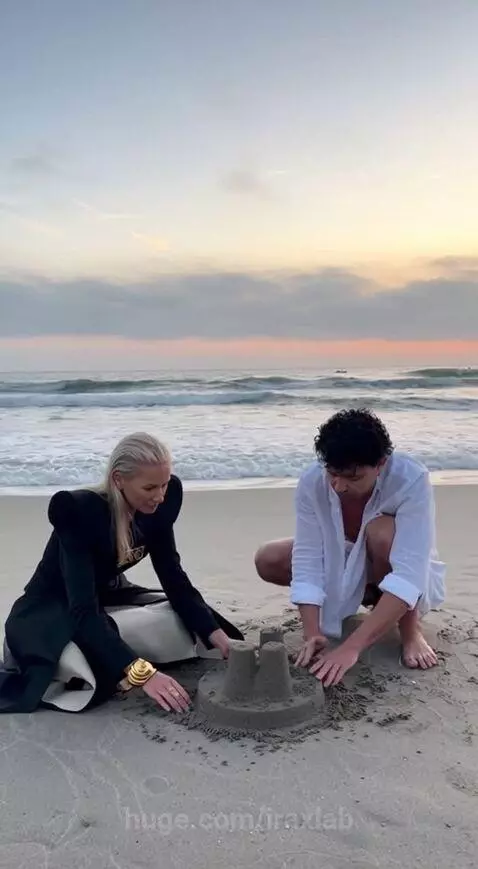 Couple building a sandcastle on a beach at sunset, with the ocean and lighthouse in the background.