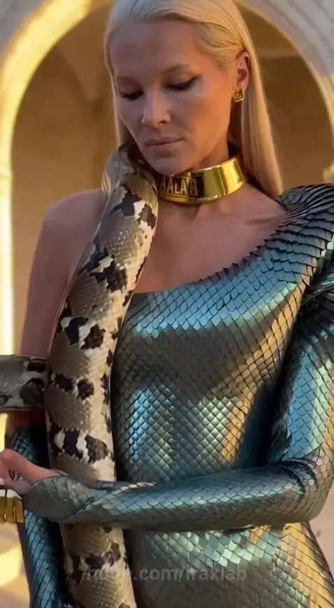 Woman in metallic green dress and black boots with a python wrapped around her neck and body in a stone setting.