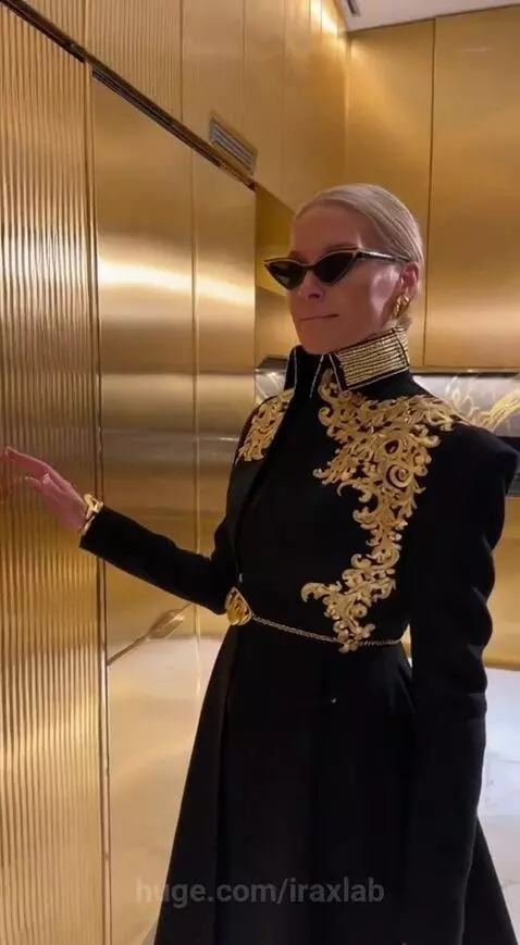 Woman in black coat with gold embroidery and sunglasses in a luxurious kitchen with gold cabinets and marble.