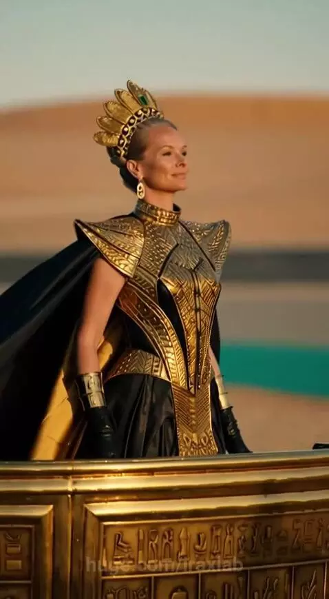 Queen of Speed in a golden costume and crown in a chariot pulled by men on a desert race track at sunset.