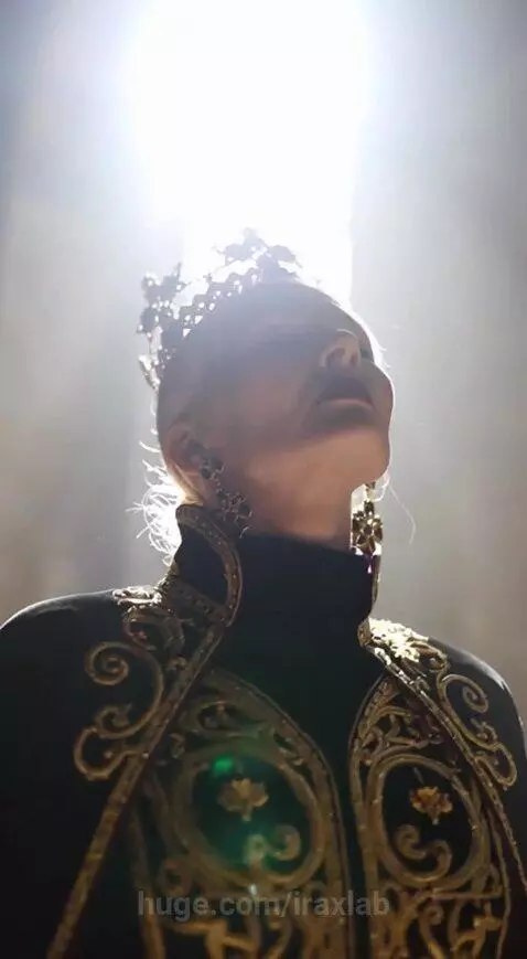 Regal queen in black gown with gold embroidery, red cape, and crown, standing in a grand hall with dramatic lighting.